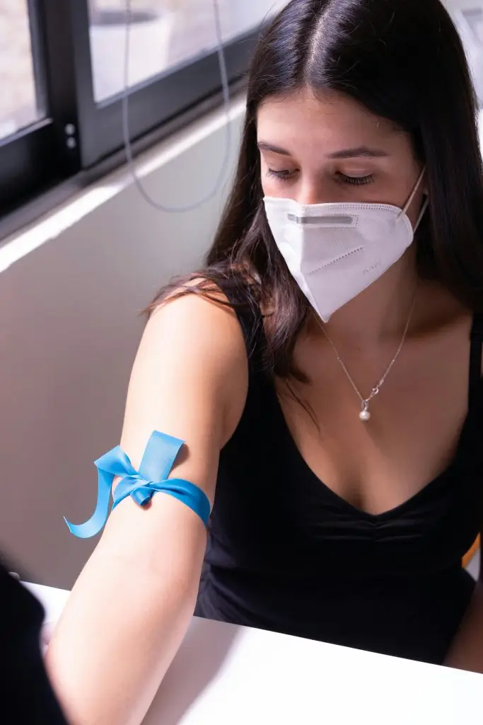 A woman wearing a face mask prepares for a blood test in a medical setting, focusing on health safety.